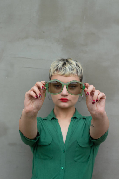 Glamorous fashion model girl with short blond hair in a green dress posing with retro green sunglasses. Bright makeup. Against the background of a concrete wall. Beauty trends. Fashion blogger - Photo, Image