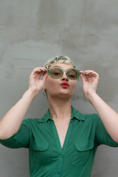 Půvabná modelová dívka s krátkými blonďatými vlasy v zelených šatech s retro zelenými slunečními brýlemi. Chytrý make-up. Na pozadí betonové zdi. Trendy krásy. Módní blogger - Fotografie, Obrázek