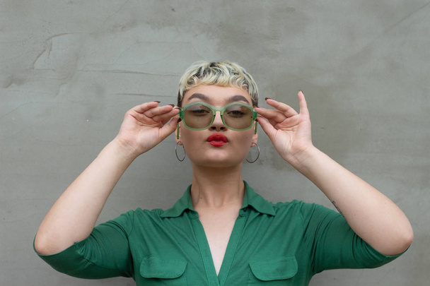 Glamouröses Modelmädchen mit kurzen blonden Haaren in einem grünen Kleid posiert mit retro-grüner Sonnenbrille. helles Make-up. vor dem Hintergrund einer Betonmauer. Beauty-Trends. Modebloggerin - Foto, Bild