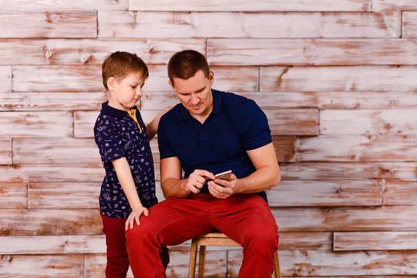 a young man shows his son a photo on a smartphone. Happy father and son with smartphone on wooden background - Foto, immagini
