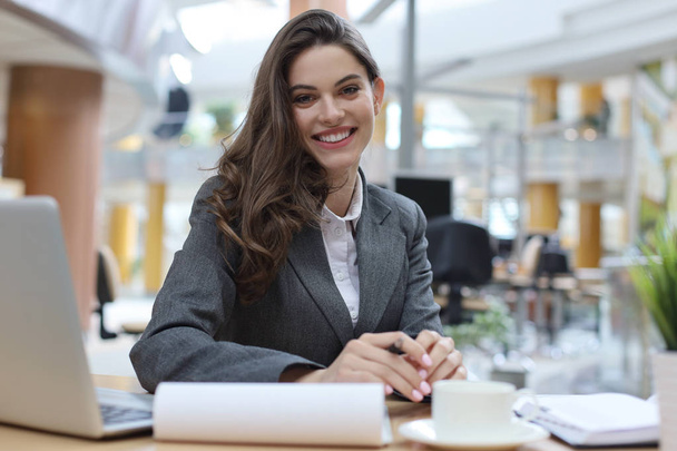 Portrait of a cheerful young businesswoman sitting at the table in office and looking at camera. - Фото, изображение