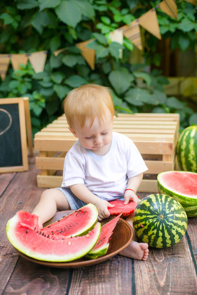 A little boy with blond hair in the summer on a sunny day sitting and eating a red tasty watermelon - Photo, Image