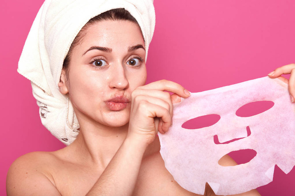 Portrait of half naked cute young woman with white towel on her head, looking directly at camera, showing face cosmetic mask after applying it, face revitalizing procedure, looking surprised. - Foto, Imagen