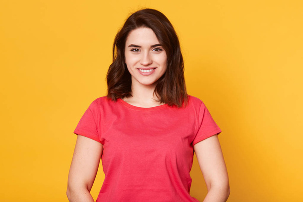 Adorable pleasant woman wearing casual red t shirt posing isolated over yellow background in studio, looking directly at camera, having sincere smile on her face, looking pleasant. Emotions concept. - Fotografie, Obrázek