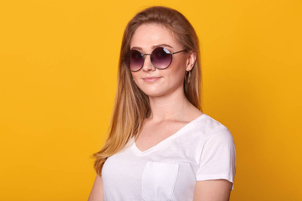 Image of sweet confident young lady standing isolated over bright yellow background in studio, wearing white casual t shirt and fashionable round eyeglasses, having long blond hair. Youth concept. - Foto, Imagem