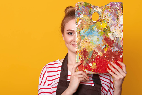 Indoor studio shot of satisfied talented girl holding palette to mix colors, covering half of her face with it, looking directly at camera, ready to paint masterpiece, being inspired by her hobby. - Photo, image