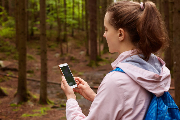 Outdoor shot of woman backpacker get lost and trying to find her way, using online maps in mobile phone in forest, female going for walk in wood. Traveling, backpacking and having adventures concept. - Foto, imagen