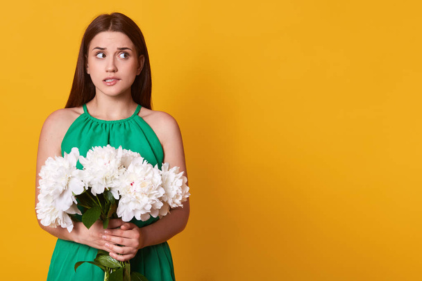 Confused worried cute model looking aside, biting her lip, standing still at one side, holding big bouquet of white peonies, having feeling of anixiety, looks nervous. Copyspace for advertisement. - Photo, image