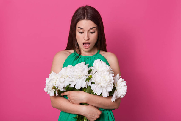 Indoor shot of shocked impressed young woman looking at bouquet with open mouth, stands embracng aring green dress. Unknown hand holding bunch of pink and white peonies. - Fotó, kép