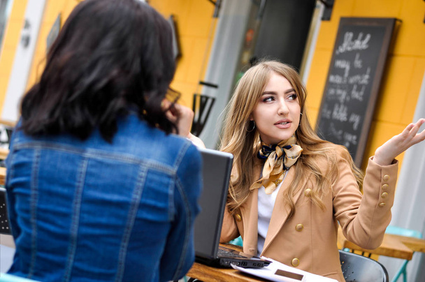 Две молодые стильные женщины сидят в городском кафе и разговаривают друг с другом
. - Фото, изображение