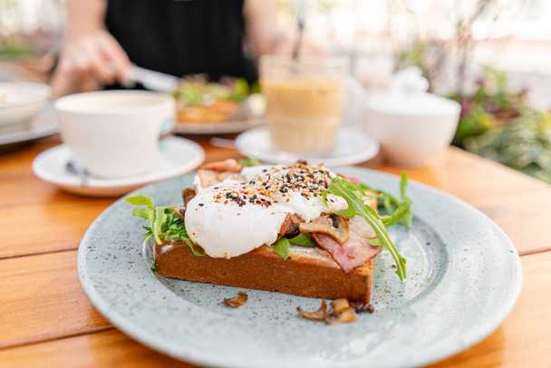 Breakfast and coffee in the summer cafe. Sandwich on a bun brioche with bacon, mushrooms, arugula and poached egg - 写真・画像