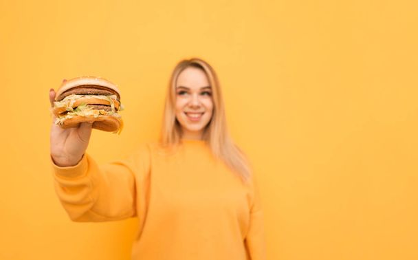 幸せな笑顔の女の子がオレンジ色のカジュアルな服を着て、黄色の背景に立って、彼の手に大きな食欲ハンバーガーのロスアップ写真。女の子の手にファーストフード。有害な食品の概念. - 写真・画像