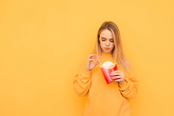 Piękna dziewczyna w pomarańczowej bluzki z frytkami w rękach wyizolowana na żółtym tle. Lady trzyma Fast food w ręku. Szkodliwa koncepcja żywności. Izolowane - Zdjęcie, obraz