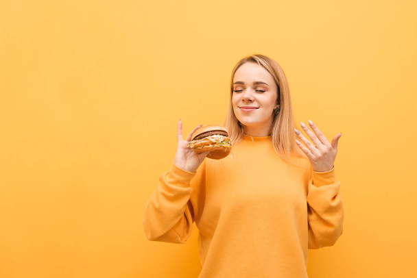 Happy girl with burger in his hands sniffs smell of fresh fast food and smiles on a yellow background. Blonde girl sniffs an appetizing burger, isolated. Fast Food Concept - Photo, Image
