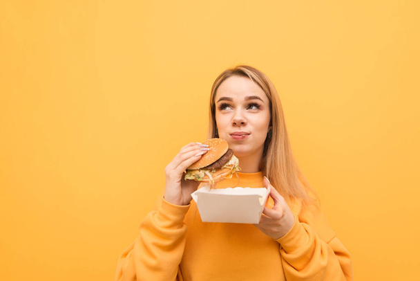 魅力的なブロンドのクローズアップポートレートは、黄色の背景に配達とハンバーガーを食べて、空白の場所を脇に見て。悪い食べ物を食べる明るい服の面白い女性 - 写真・画像