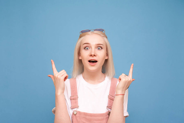 Portrait teenage girl with light hair, looks at the camera with astonishment and shows fingers up on the ad.Funny girl in casual dress with a surprised face, showing fingers up. Isolated - Photo, Image