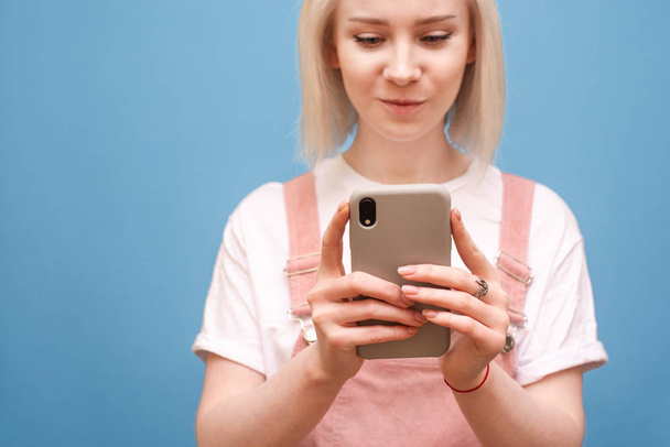 Okostelefon a kezében egy tini lány. Közeli kép egy szőke lány aranyos ruhát használ egy okostelefon a kék háttér, a hangsúly a telefon a kezében a lány. Copyspace - Fotó, kép
