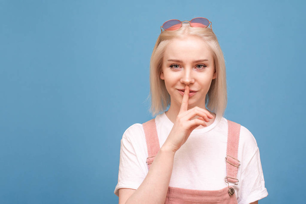 A portrait of a funny teen girl shows a silence sign on a blue background and looks at the camera. Funny girl isolated on a scarlet background, joined the index finger to the lips. Copyspace - Photo, Image
