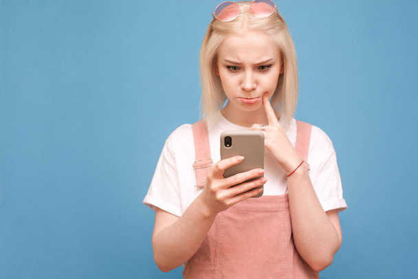 Legrační blondýnka v roztomilým oblečení s zamyšlenou tváří používá smartphone na modrém pozadí, soustředění se na telefon v rukou dívky. Copyspace - Fotografie, Obrázek