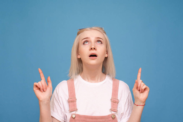 Emotional girl in a bright casual dress looks and shows fingers up on a blank space, isolated on a blue background. Surprised teenage girl shows fingers up on copyspace - Photo, image