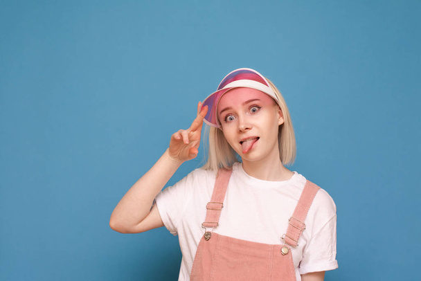 Portret zabawnej dziewczyny w dość przypadkowym stroju, ubrany w różową czapkę i zrobienie śmiesznego oblicza na niebieskim tle. Zabawna dziewczyna pozuje na kamerę, wyizolowany portret zbliżenie. - Zdjęcie, obraz
