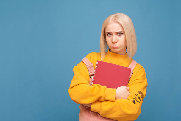 Portrait of a girl's funny student stands on a blue background, keeps books and books in her hands and looks sadly in the camera. Disappointed teenage girl poses on the camera with books in his hands - Fotografie, Obrázek