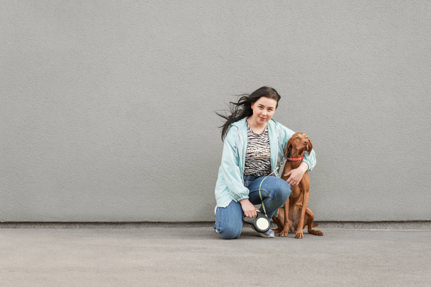 Happy woman sits with a dog and hugs a gray wall against the background, looks at the camera and smiles. Owner poses with the dog against the background of the wall. Copyspace - Photo, Image