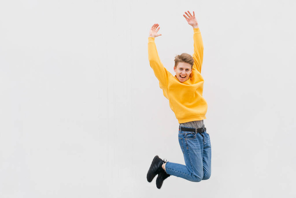 Jump of a happy young teenager in a casual dress up against a white wall, wearing jeans, yellow sweatshirt and sneakers. Joyful student jumping against the background of a white wall, street portrait - Photo, image
