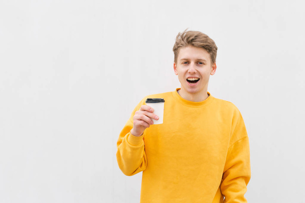 fröhlicher Typ mit einer Tasse Kaffee auf dem Hintergrund einer weißen Wand, der in die Kamera blickt. ein ausdrucksstarker junger Mann hält einen Pappbecher in der Hand, isoliert vor dem Hintergrund einer hellen Wand. - Foto, Bild