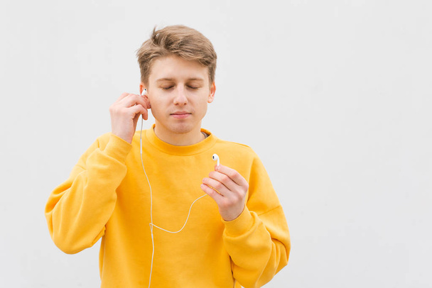 Closeup portrait of a young man stands on the background of a white wall,inserts a wired headset in his ears with his eyes closed and enjoys music.Guy with headphones on the background of a white wall - Fotoğraf, Görsel