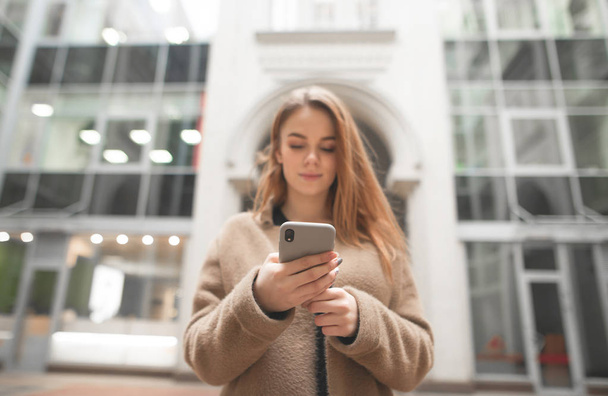 Stilvolle Mädchen nutzt ein Smartphone auf der Straße, konzentriert sich auf den Handybildschirm, vor dem Hintergrund der modernen Architektur, den Fokus auf das Smartphone. Mädchen schreibt eine Nachricht am Telefon. - Foto, Bild