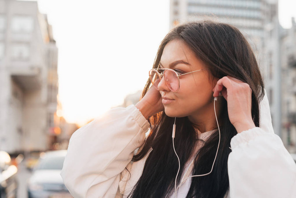 Close-up street portrait of a brunette woman wearing glasses and a white jacket, headphones standing at the background of the sunset, looking sideways. Trendy girl walking along the street - Foto, Imagen