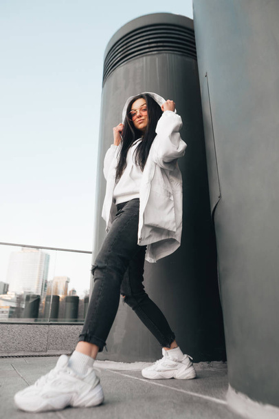 Fashionable street portrait of a girl in casual clothing, wearing a white jacket, sneakers and glasses, stands in full height, poses and looks at the camera. Street fashion. Vertical photo. - Фото, изображение