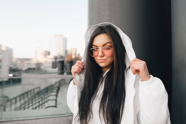 Stylish attractive girl in casual clothing, wearing pink glasses and a white jacket, posing against the background of the city landscape, looking into the camera. Street fashion portrait - Foto, imagen