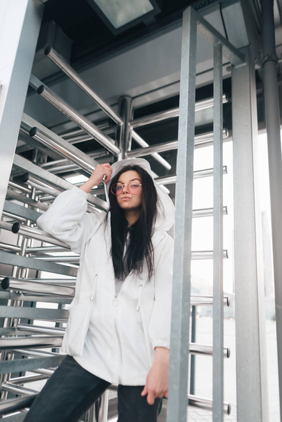 Street portrait of an attractive girl in street clothes near turnstiles. Stylish girl model in pink glasses and a white jacket poses for a futuristic location. Street fashion concept. - Photo, Image