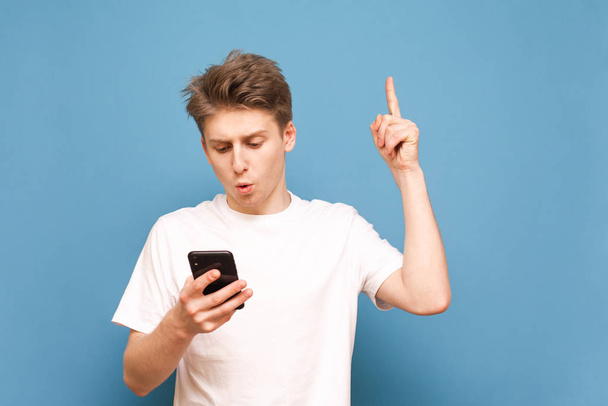 Хлопець у білій футболці тримає смартфон в руках, він дивиться на екран з здивованими очима і показує пальцем до пустого простору, ізольованим на синьому фоні. Copyspace - Фото, зображення