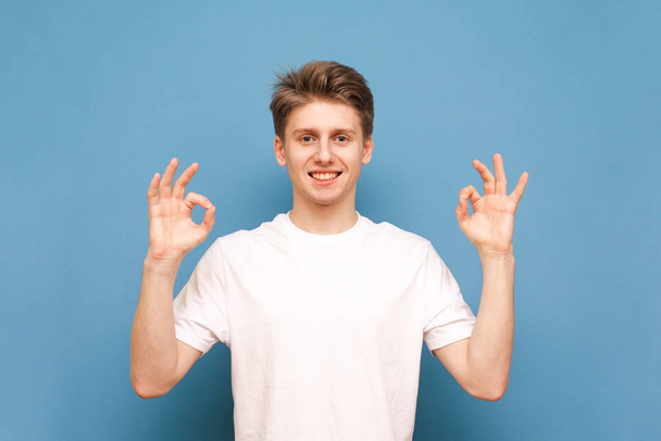 Glimlachende vent in een wit T-shirt toont een OK gebaar met zijn handen en kijkt naar de camera op een blauwe achtergrond. Happy Young Man toont een OK-teken, geïsoleerd. - Foto, afbeelding