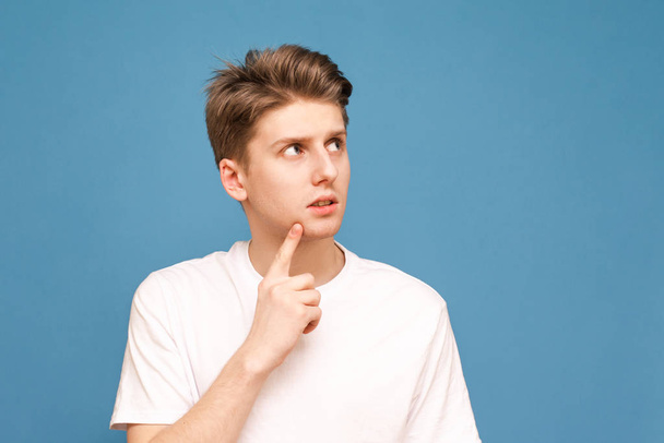 Portrait of a guy in a white T-shirt stands on a blue background, looks aside and thinks. Thoughtful young man poses on a blue background and looks at a place for advertising. Isolated. - Foto, Bild