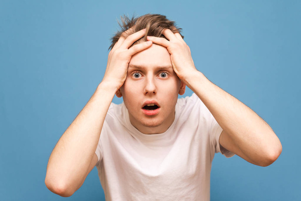 Portrait of a shocked guy on a blue background. Young man in a white shirt took his hands behind his head and emotionally looks into the camera, looks nervously into camera, demonstrates phobia - Photo, Image