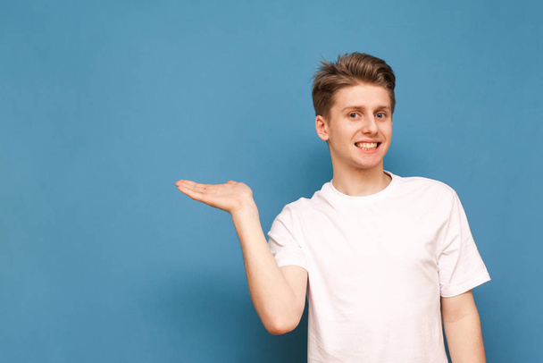 Grappige jonge man in een wit T-shirt staat op een blauwe achtergrond en toont zijn hand in de plaats voor de tekst, kijkt naar de camera en glimlacht. Geïsoleerd. - Foto, afbeelding