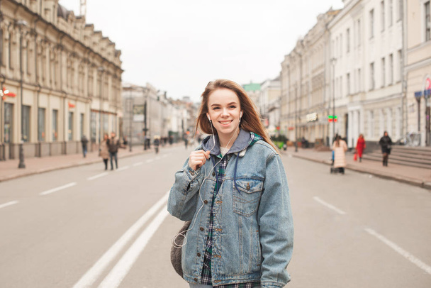 ヘッドフォンを耳にして街を歩き回り、音楽を聴き、笑顔でジーンズジャケットを着て街を歩く幸せな少女の肖像画。通りに立って微笑む学生の女の子 - 写真・画像