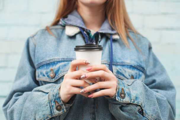 A menina de foto close-up em uma jaqueta de ganga mantém uma xícara de papel com café no fundo de uma parede azul. Fotografia disparada. Espaço de cópia
 - Foto, Imagem