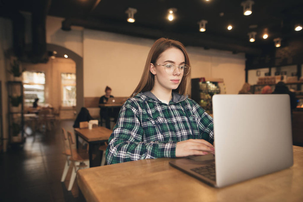 Привлекательная девушка в очках, одетая в рубашку, использует ноутбук в уютном кафе, сосредоточена на просмотре экрана. Студентка учится с ноутбуком в кофейне
. - Фото, изображение
