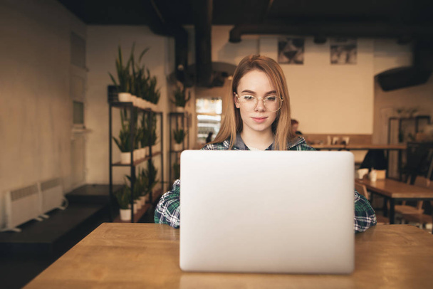 Piękna dziewczyna freelance pracuje w przytulnej kawiarni na laptopie. Uczennica w koszulce studiujących w kawiarni, korzystając z Internetu na laptopie. Praca w Internecie. - Zdjęcie, obraz
