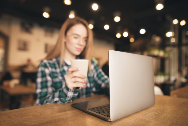 Background. Young girl works on a laptop in a cafe and holds a glass of coffee in her hand. Woman uses a laptop in the coffee shop. Focus on the laptop. - 写真・画像