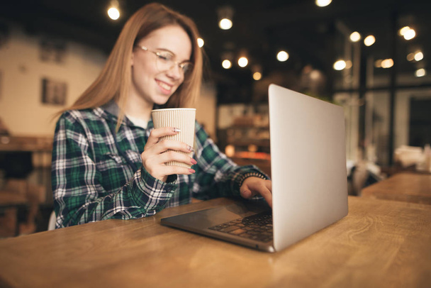 Щаслива дівчина зі склянкою кави в руках використовує ноутбук у кафе, дивиться на екран і посміхається. Фокус на склянці кави в руці
. - Фото, зображення