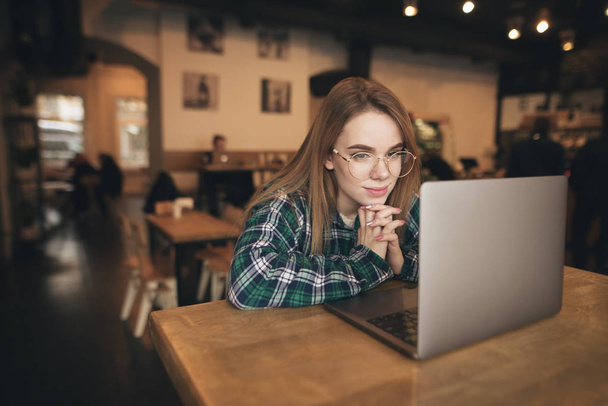 Portret uśmiechniętej dziewczyny w casual ubrania i okulary siedzą w przytulnej kawiarni z laptopem, stwarzając na aparacie. Happy Girl korzysta z laptopa w kawiarni i patrzy na kamerę - Zdjęcie, obraz