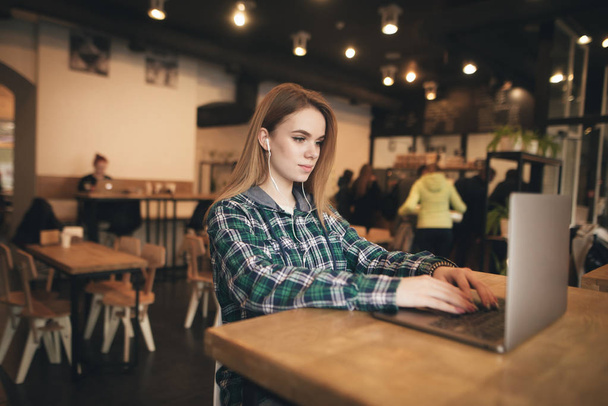 Retrato de uma menina elegante vestindo uma camisa, usando um laptop no café, olhando para a tela e ouvindo o som nos fones de ouvido. Menina bonita trabalha em um laptop em um café
. - Foto, Imagem