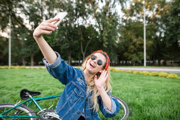 Felice bella ragazza in abiti eleganti e occhiali da sole si siede sull'erba nel parco e prende selfie sullo sfondo bike.Young ragazza si siede nell'erba nel parco e prende selfie su uno smartphone
 - Foto, immagini