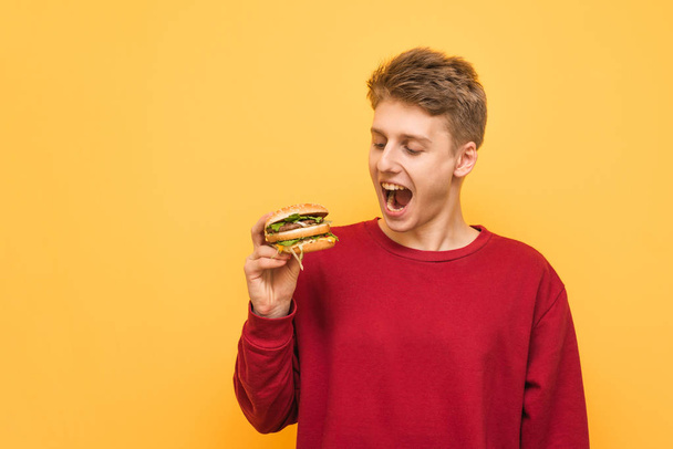 Verraste jonge man kijkt naar de hamburger in zijn handen, geïsoleerd op een gele achtergrond. Portret van emotionele man op gele achtergrond met hamburger in zijn handen. Fast Food. - Foto, afbeelding